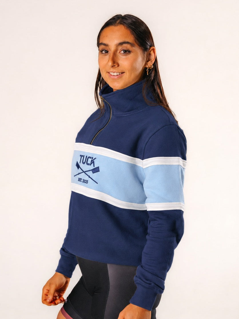 A woman modeling a navy blue quarter-zip sweatshirt with a light blue and white chest stripe featuring the 'TUCK' brand logo, paired with black athletic shorts, against a plain white studio background.